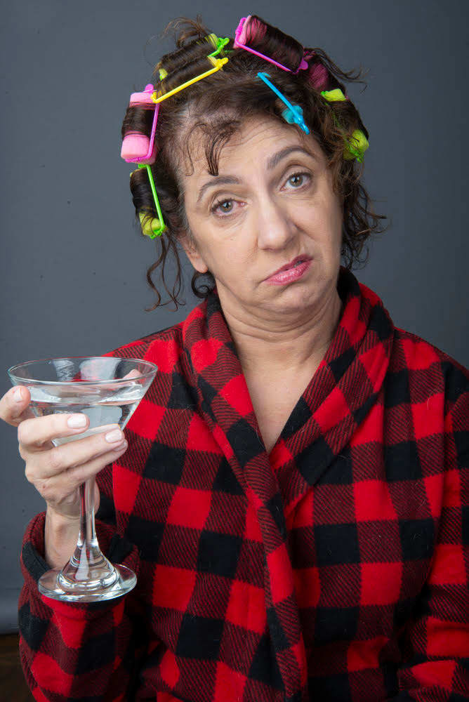 Nancy Levine in character as the Bad Substitute in a red plaid bathrobe with colorful curlers and a martini