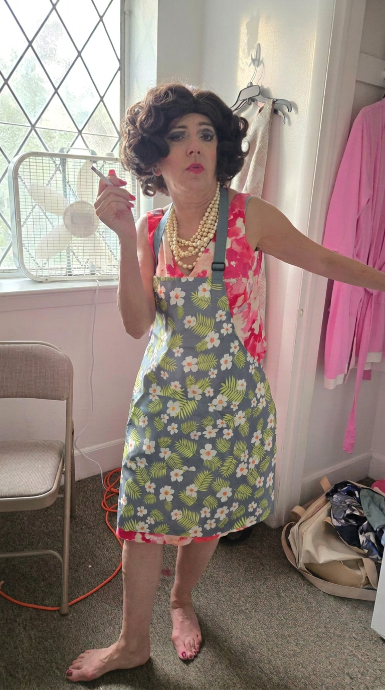 Nancy in a floral apron and pearls holding a cigarette with a surprised expression