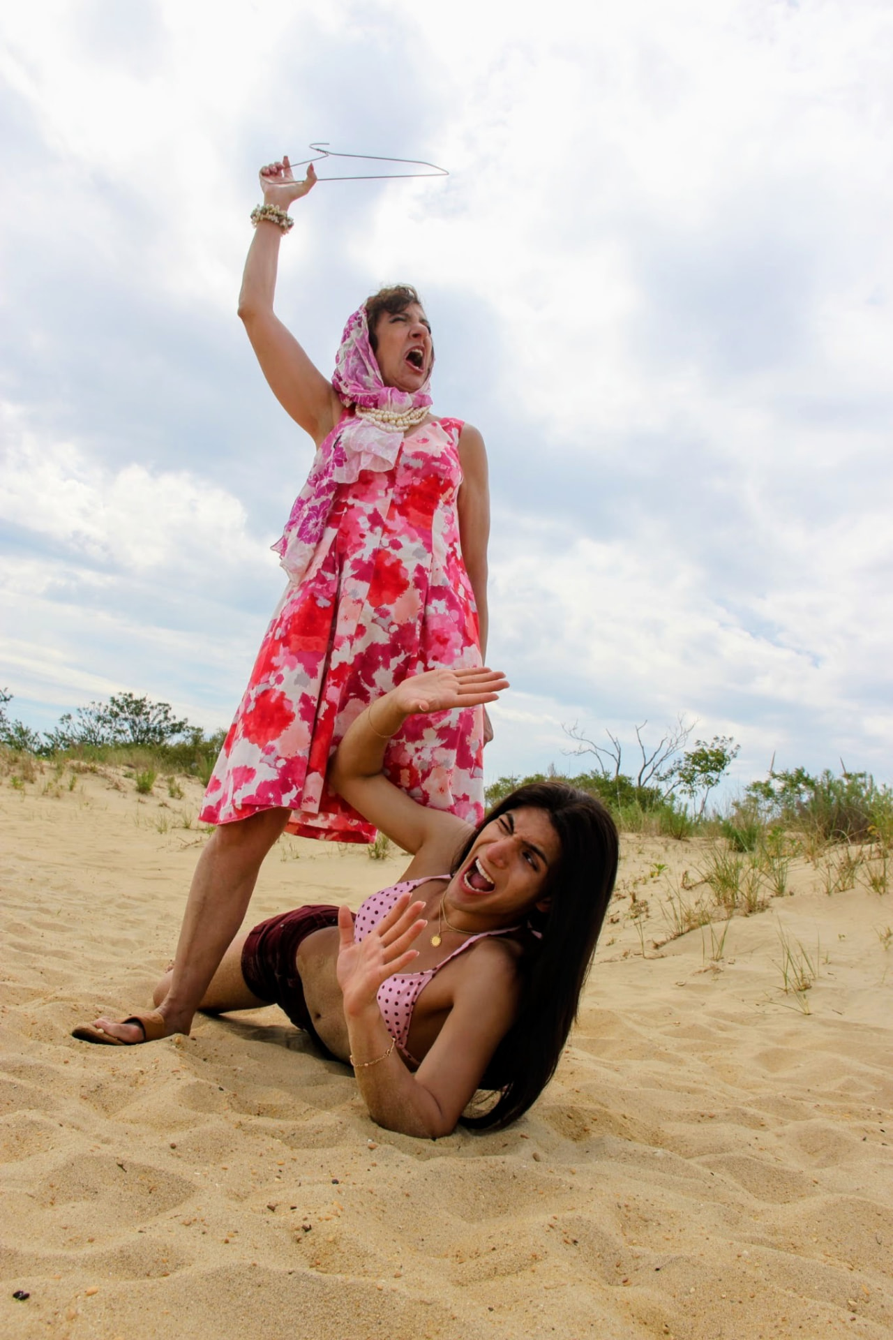 Nancy on the beach in a pink floral dress brandishing a wire hanger over a co-star lying in the sand