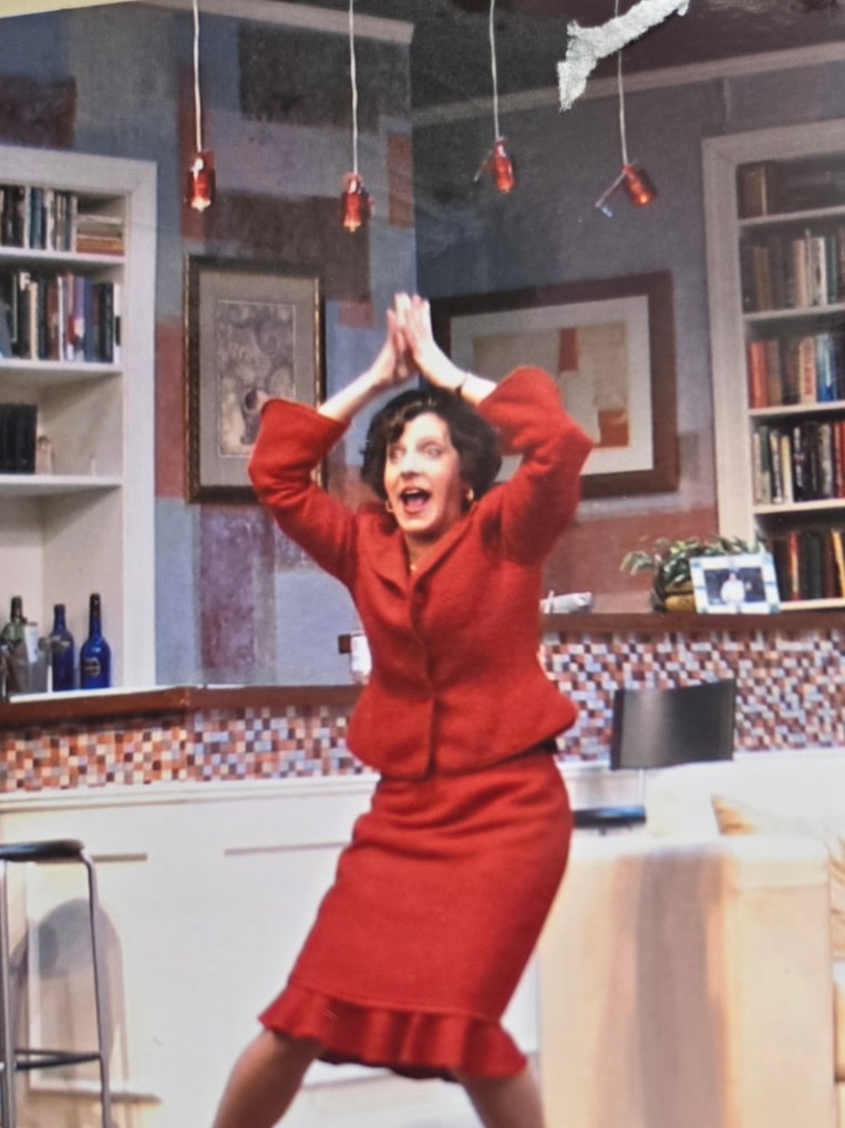 Nancy in a red suit dancing with arms overhead in a living room set