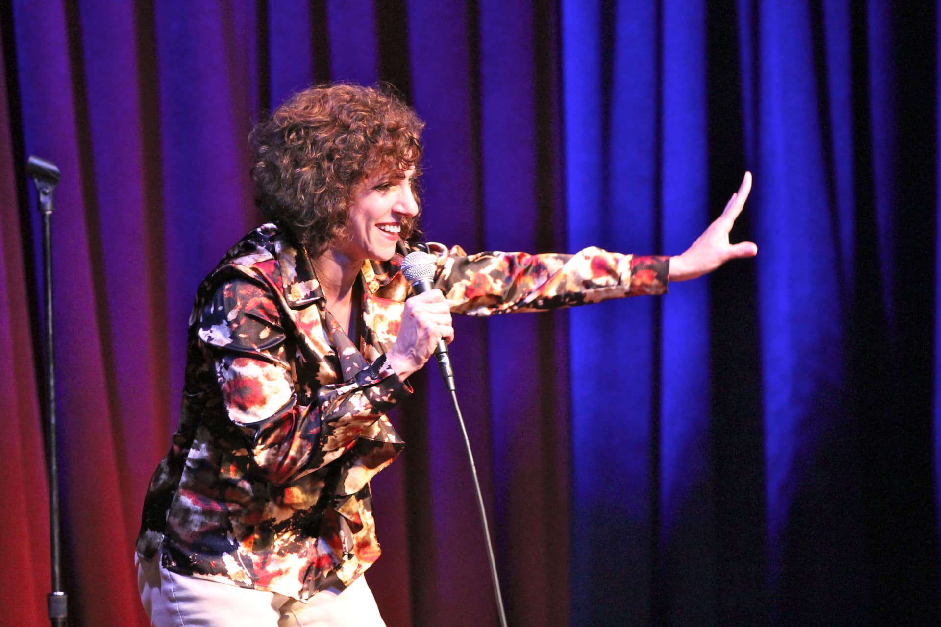 Nancy performing stand-up at Levity Live in a floral shirt with blue and purple curtains